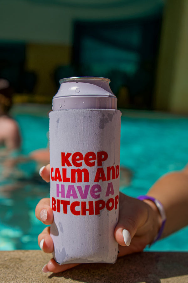 Hand holding a seltzer can with 'Keep Calm and Have a Bitchpop' koozie by a pool